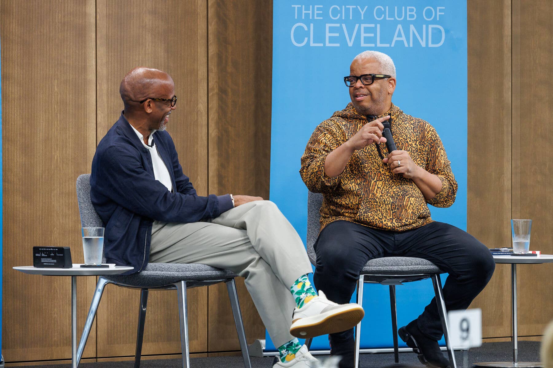 Two men sit on stage panels, one speaking into a microphone, with The City Club of Cleveland backdrop behind them