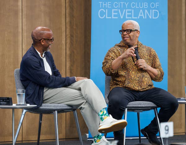 Two men sit on stage panels, one speaking into a microphone, with The City Club of Cleveland backdrop behind them