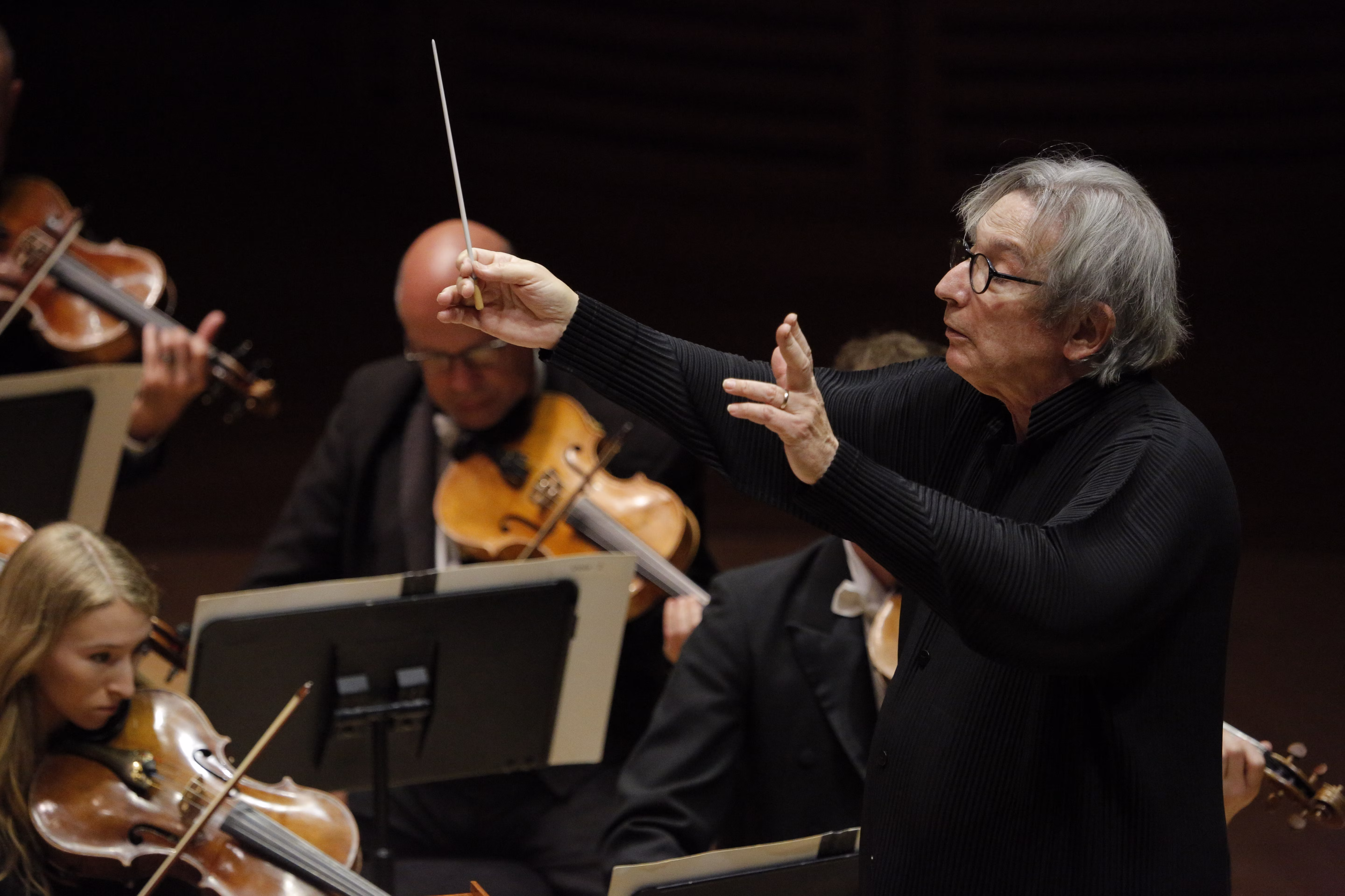 A man in a black shirt and glasses conducts an orchestra