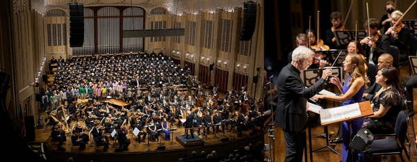 An interior shot of a concert hall showing a large orchestra and chorus onstage. A conductor standing in front of a group of singers.