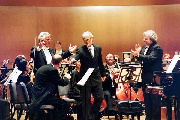 Three men in suits stand onstage with musicians seated behind them.