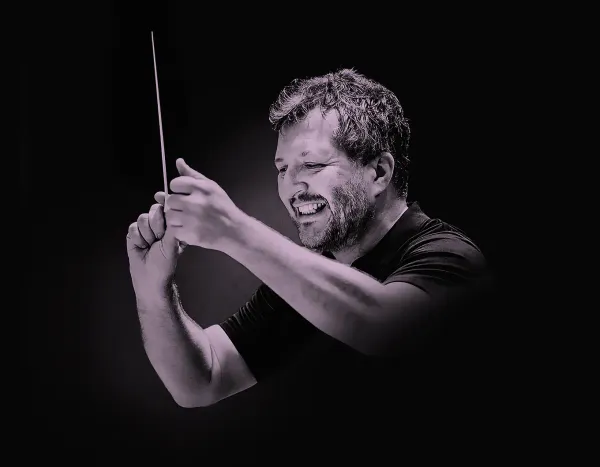 Thomas Adès smiling while conducting with a baton against a dark background.