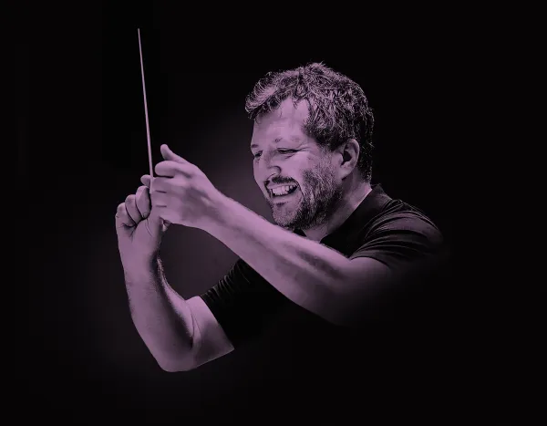 Thomas Adès smiling while conducting with a baton against a dark background.