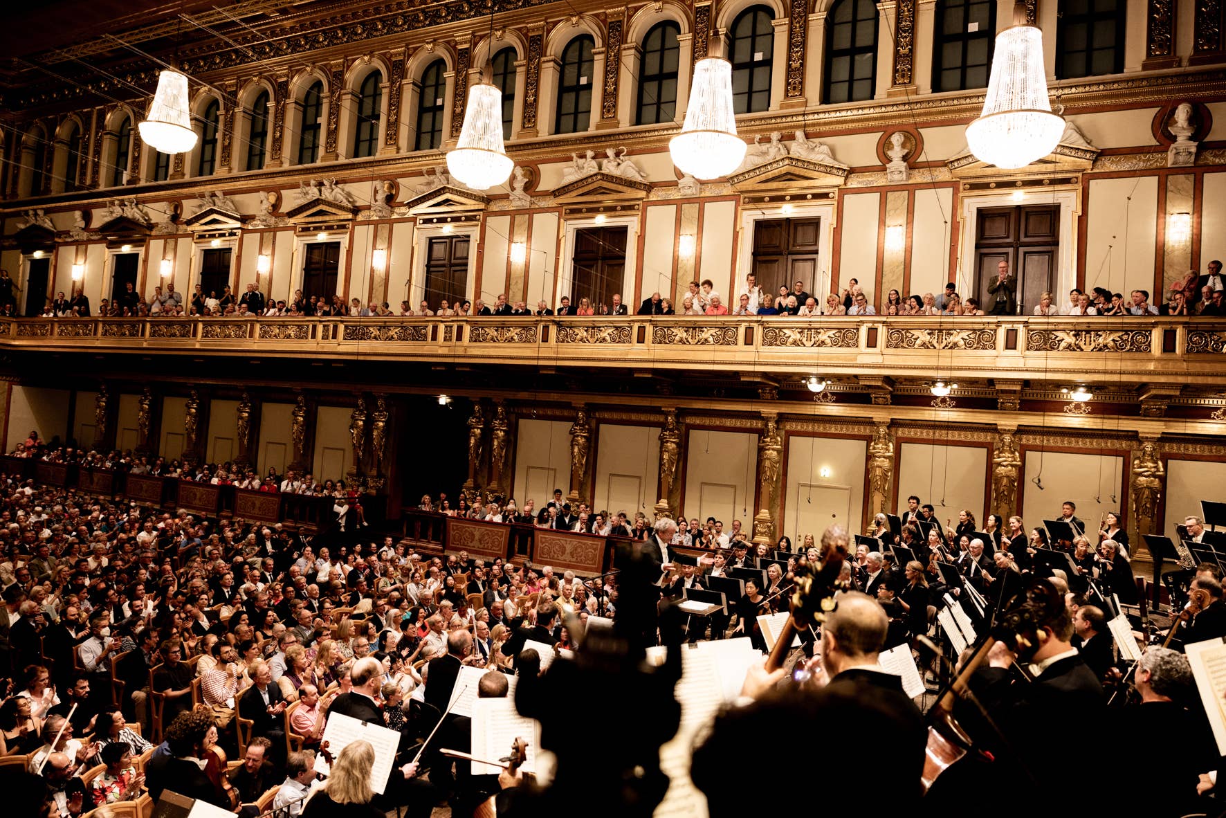 Orchestra performing in an ornate concert hall with a large audience seated and standing on balconies.