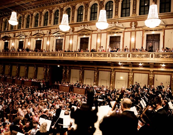 Orchestra performing in an ornate concert hall with a large audience seated and standing on balconies.