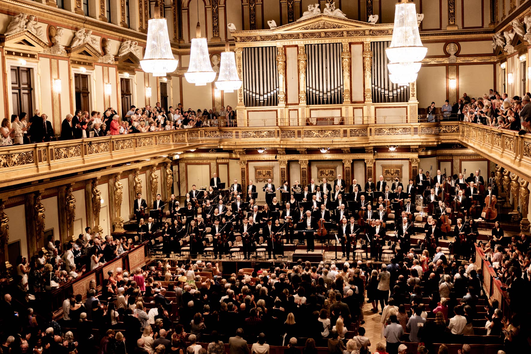 Orchestra and conductor receive a standing ovation in an ornate concert hall with chandeliers and a large audience.