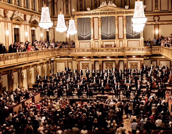 Orchestra and conductor receive a standing ovation in an ornate concert hall with chandeliers and a large audience.