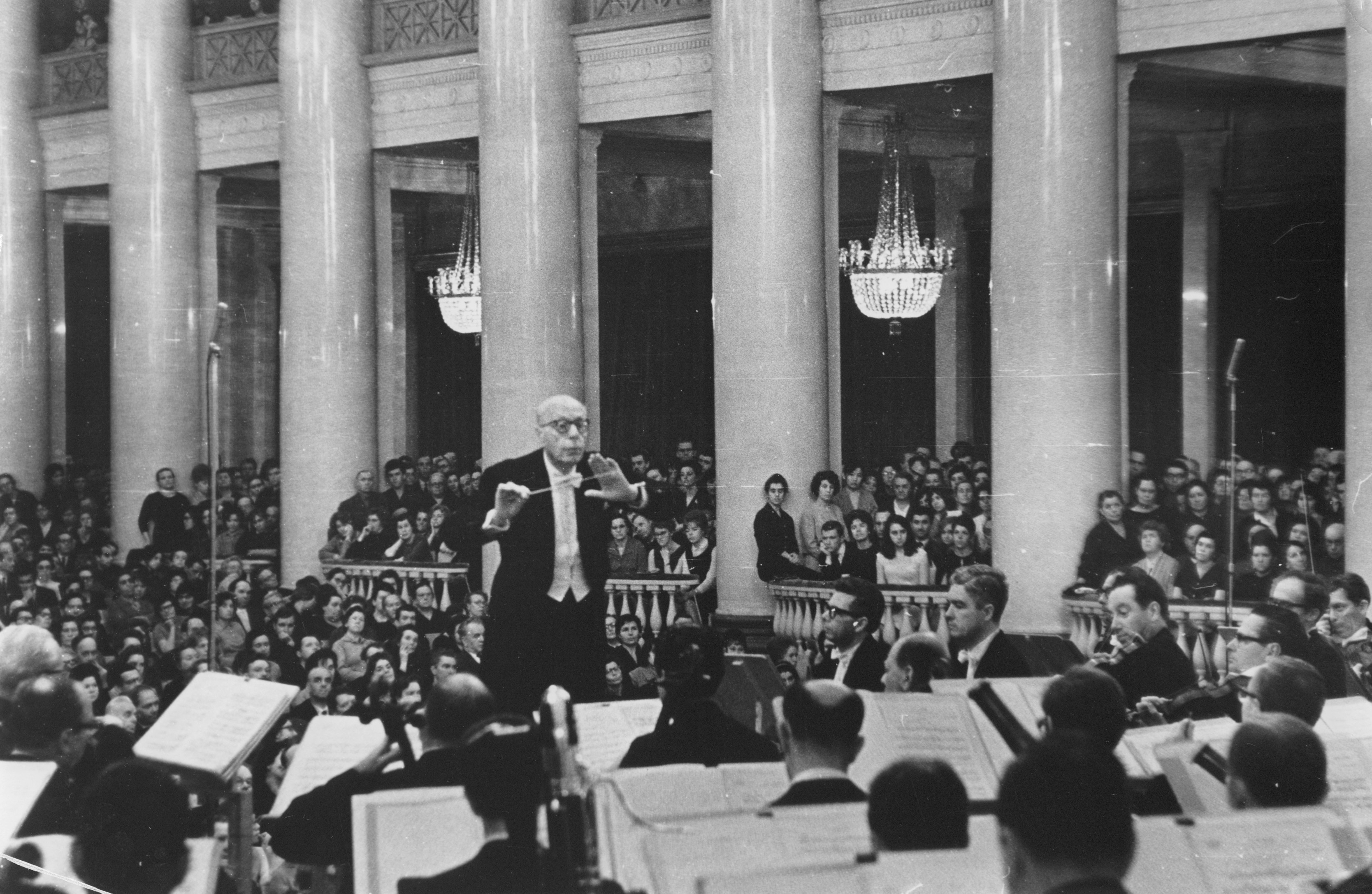 Szell is conducting the Orchestra. A large, attentive audience is seated behind them. Prominent chandeliers hang above the audience.