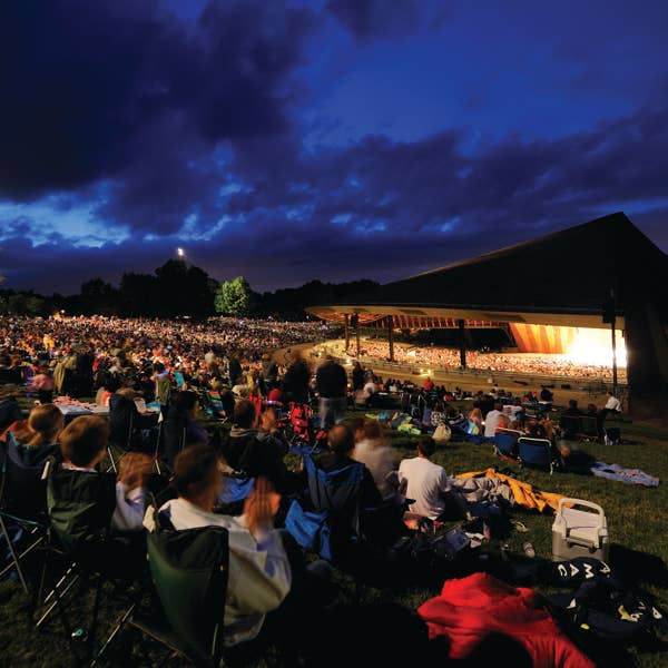under a dark blue sky, people in lawn chairs look forward to an illuminated stage