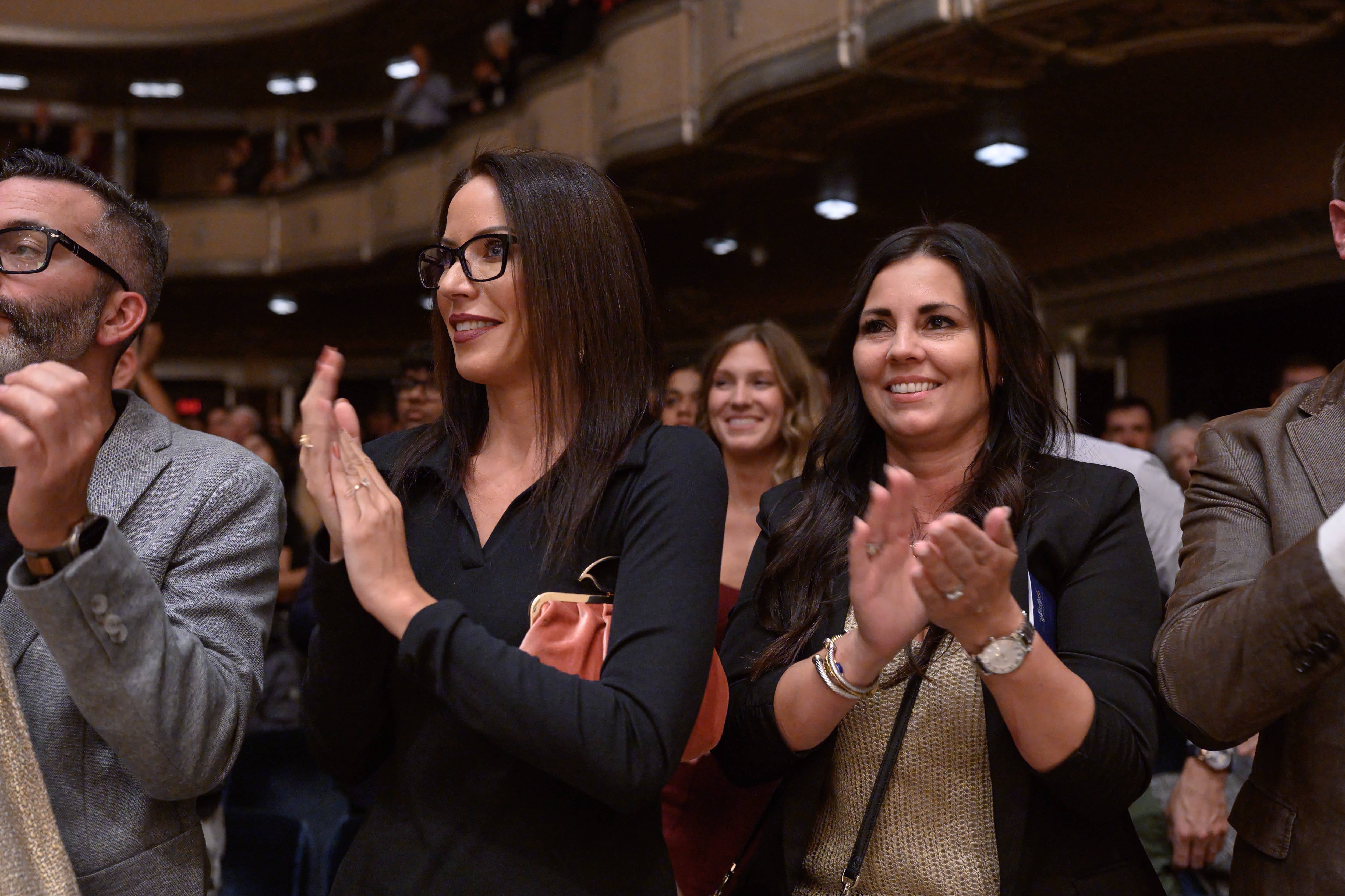 Women in business attire applauding in an auditorium with a blurred audience behind them