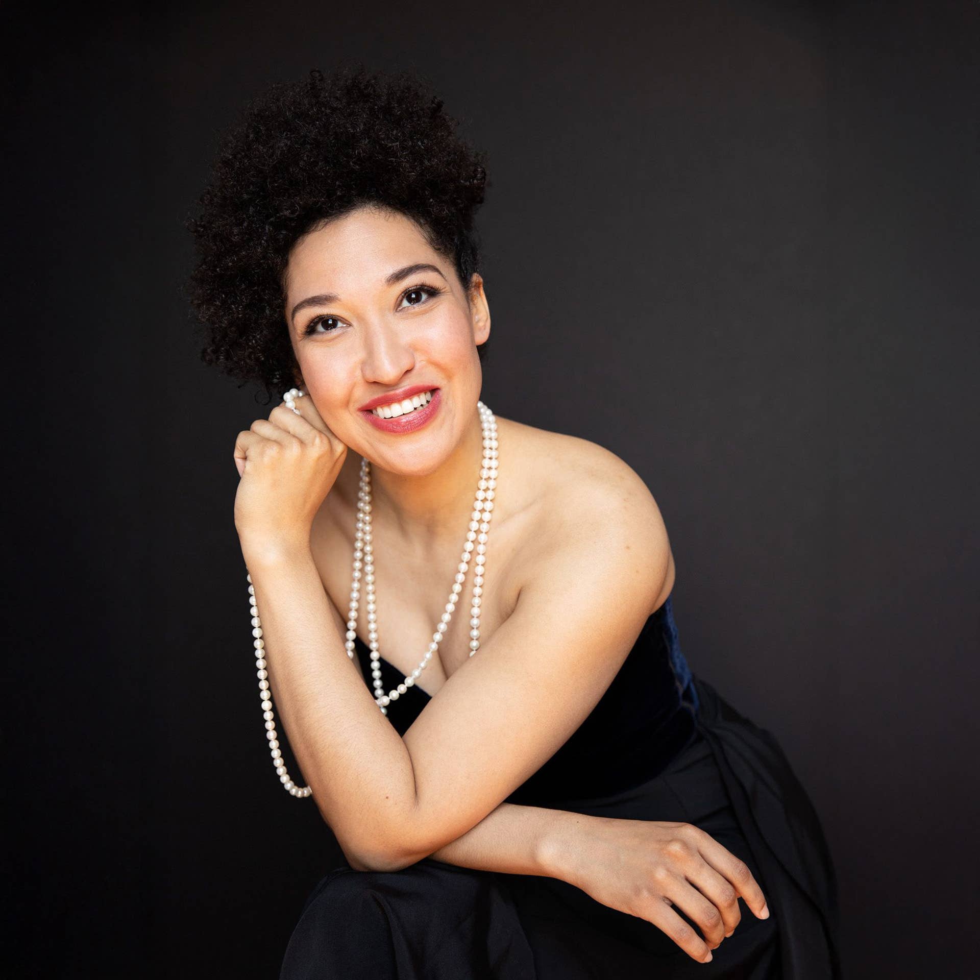 Julia Bullock posing against a dark background in a black dress with pearl necklaces