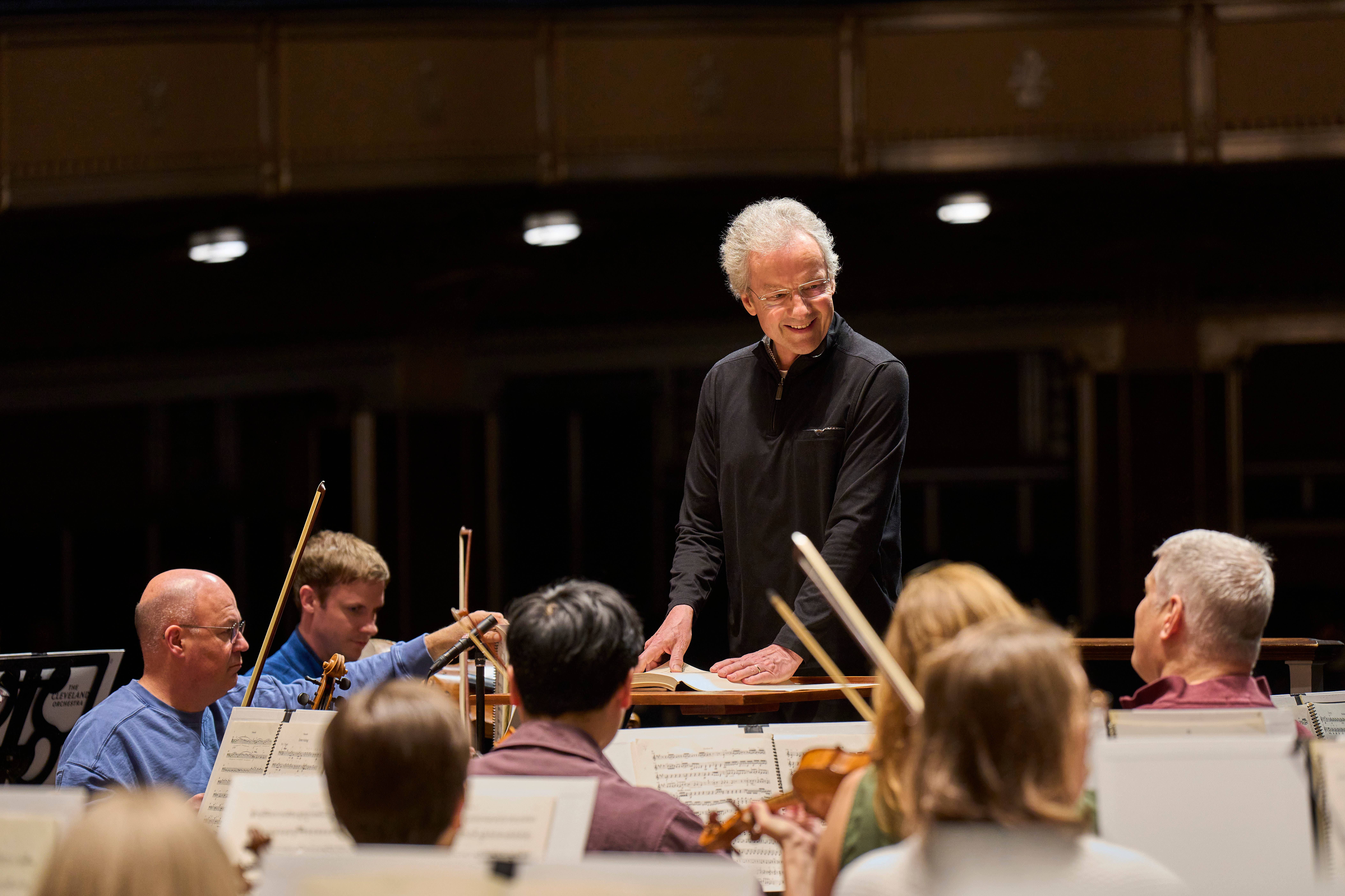 Franz Welser-Möst stands at orchestra rehearsal, smiling as musicians play with sheet music and instruments on stage.