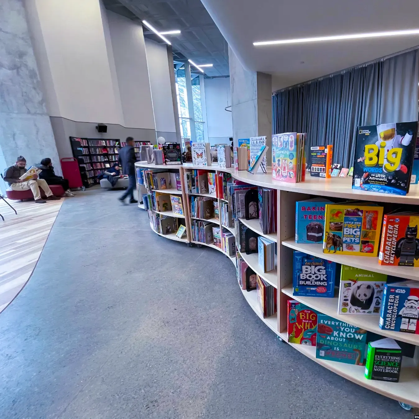 Bright library reading area with curved book display shelves, colorful children’s book covers, and seated patrons.