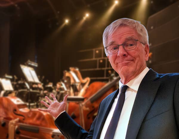 Rick Steves speaks with hand raised in an auditorium with orange seating.