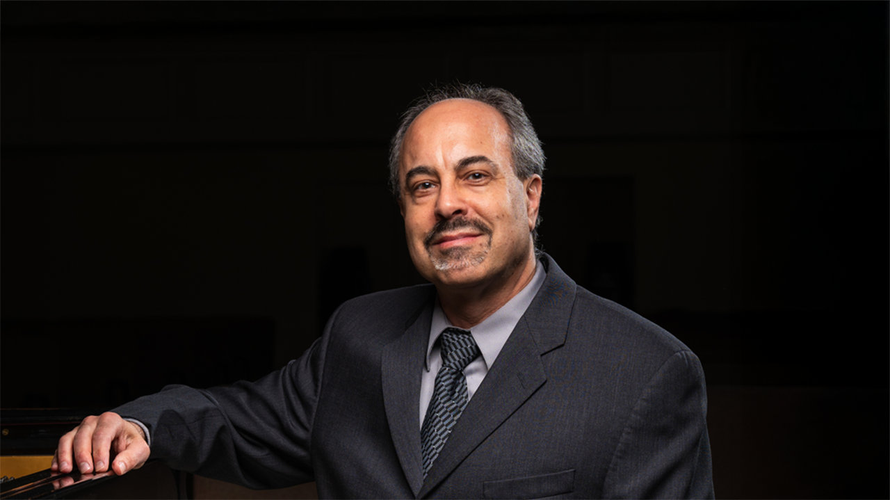 a man in a grey suit sits with his arm on a piano