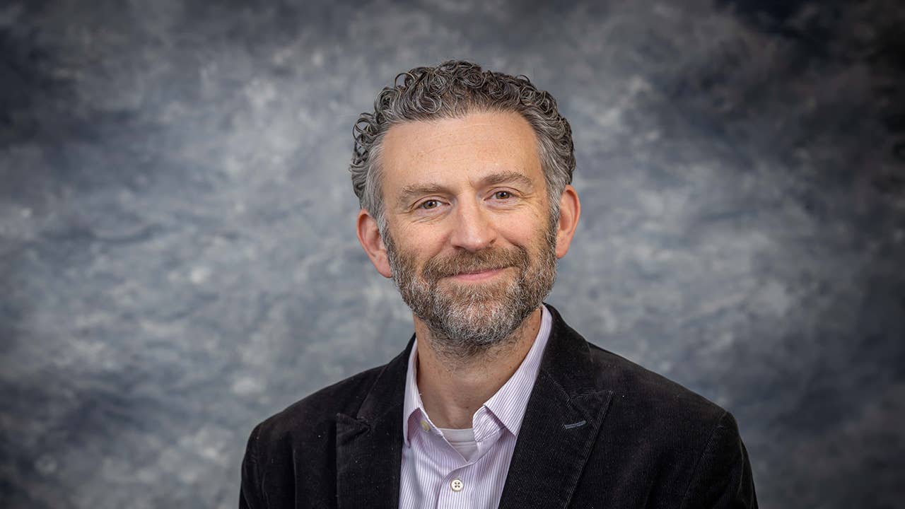 A man in a striped shirt and sports jacket smiles at the camera