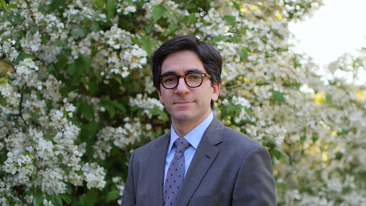 A man with black hair and glasses wears a grey suit jacket in front of white flowers