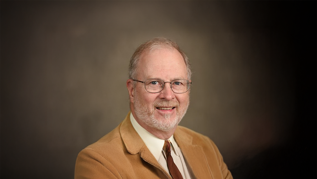 An older man in a tan corduroy jacket smiles at the camera
