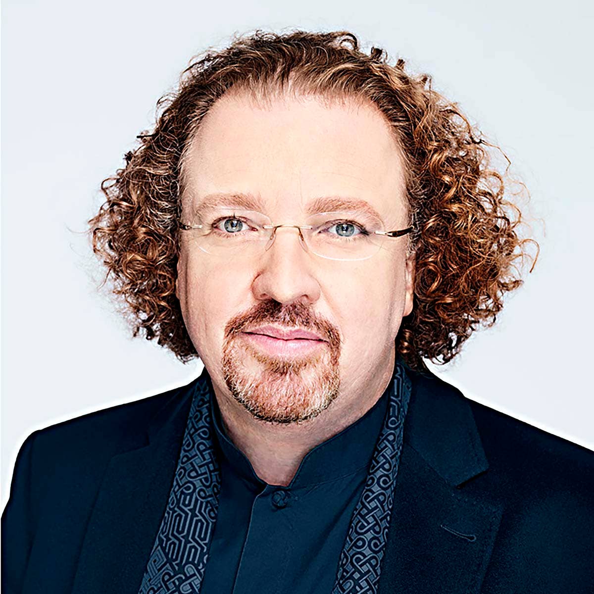 Stéphane Denève with curly hair, glasses, and a goatee, wearing a dark jacket and patterned scarf, against a light background.