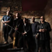 Five men pose on a brick staircase in dark suits, one wearing sunglasses, with warm spotlight lighting.