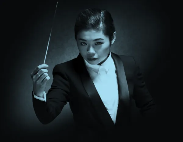 Conductor, Elim Chan, in a dark suit holding a baton, poised against a dark background with dramatic lighting.