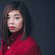 Elim Chan with straight brown hair wearing a red coat, looking at the camera against a dark background.