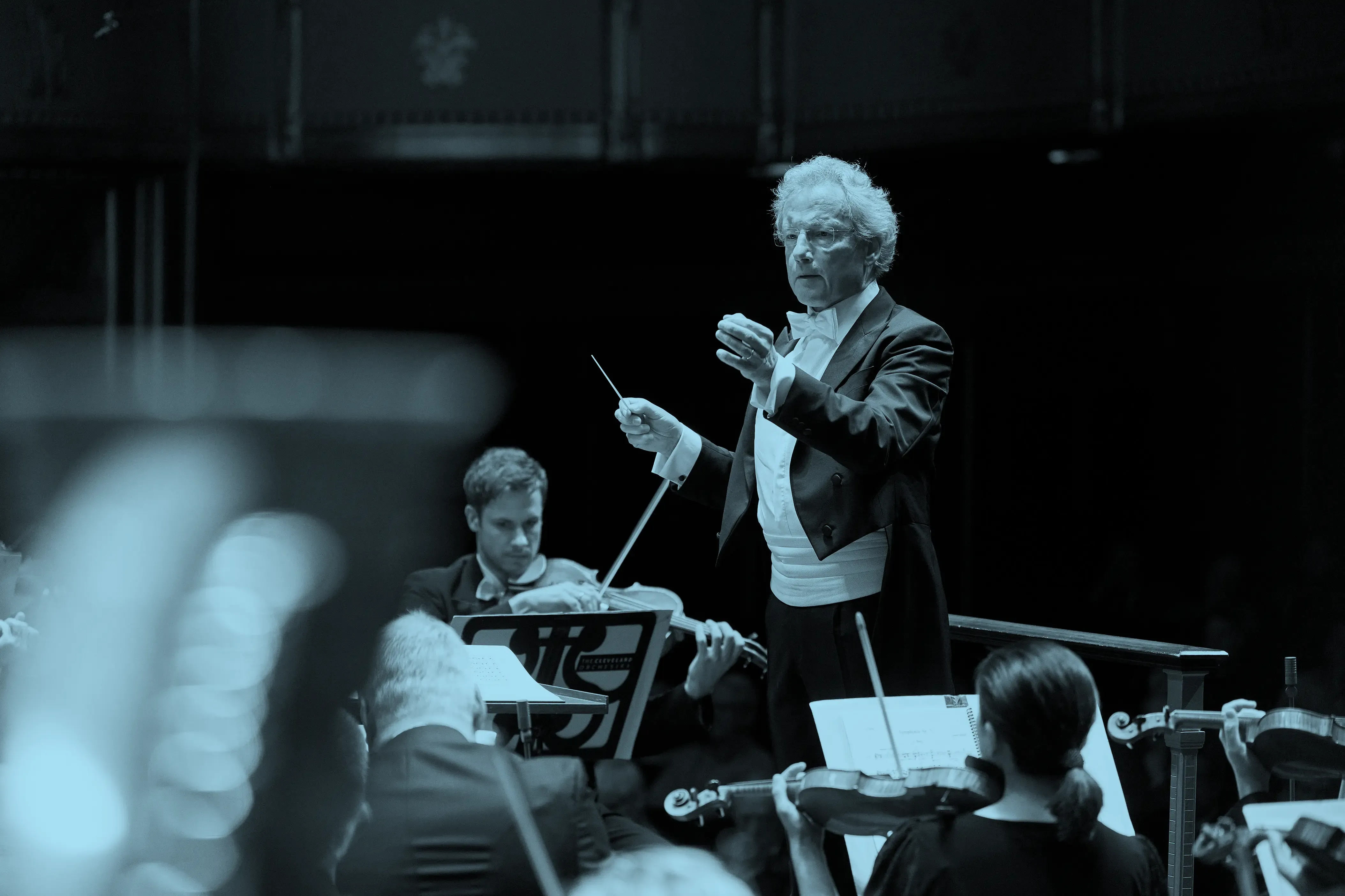 Franz Welser-Möst leading the orchestra with a baton, musicians playing string instruments, and sheet music visible.