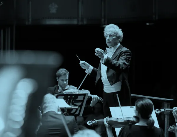 Franz Welser-Möst leading the orchestra with a baton, musicians playing string instruments, and sheet music visible.