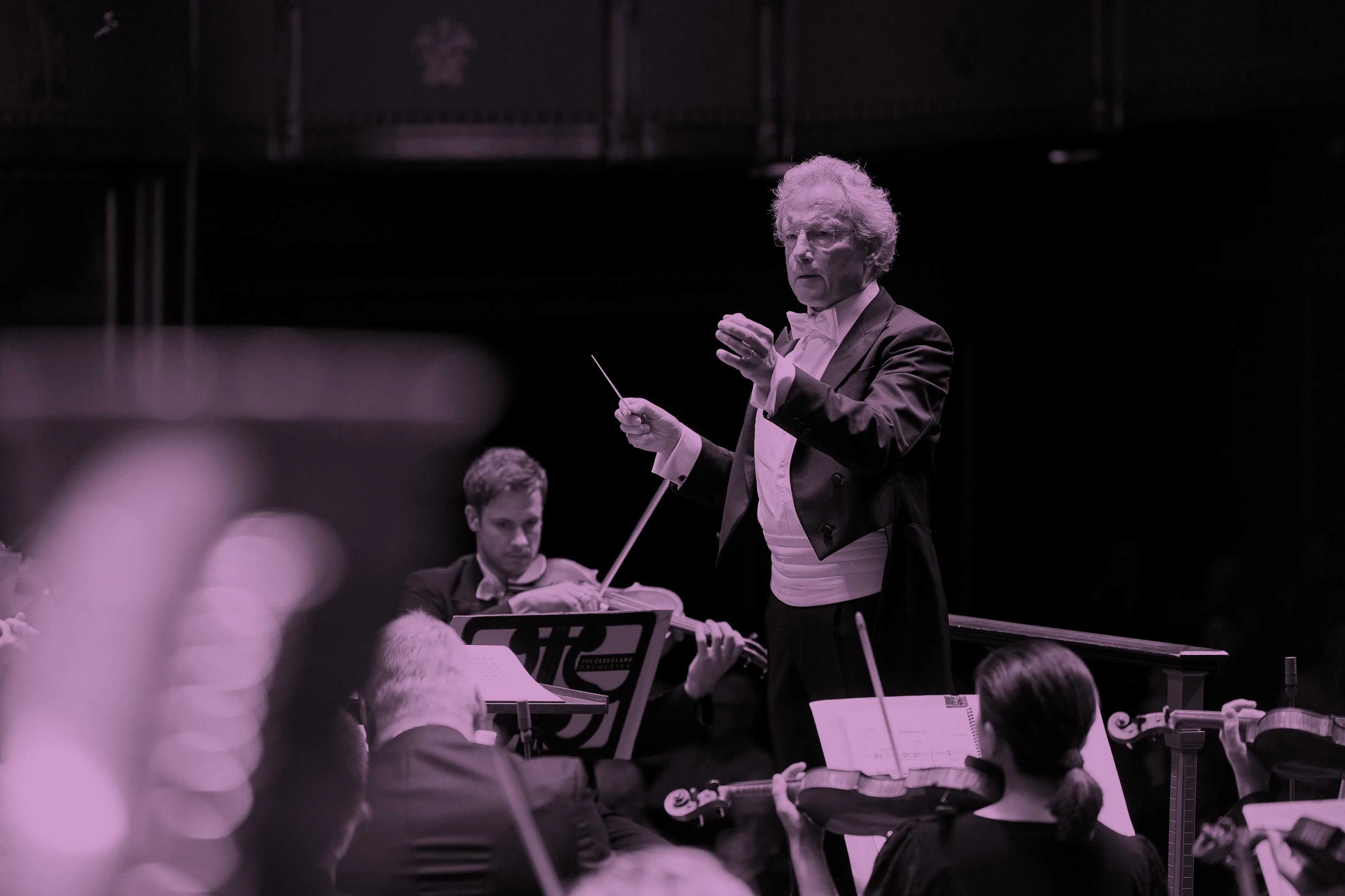 Franz Welser-Möst leading the orchestra with a baton, musicians playing string instruments, and sheet music visible.