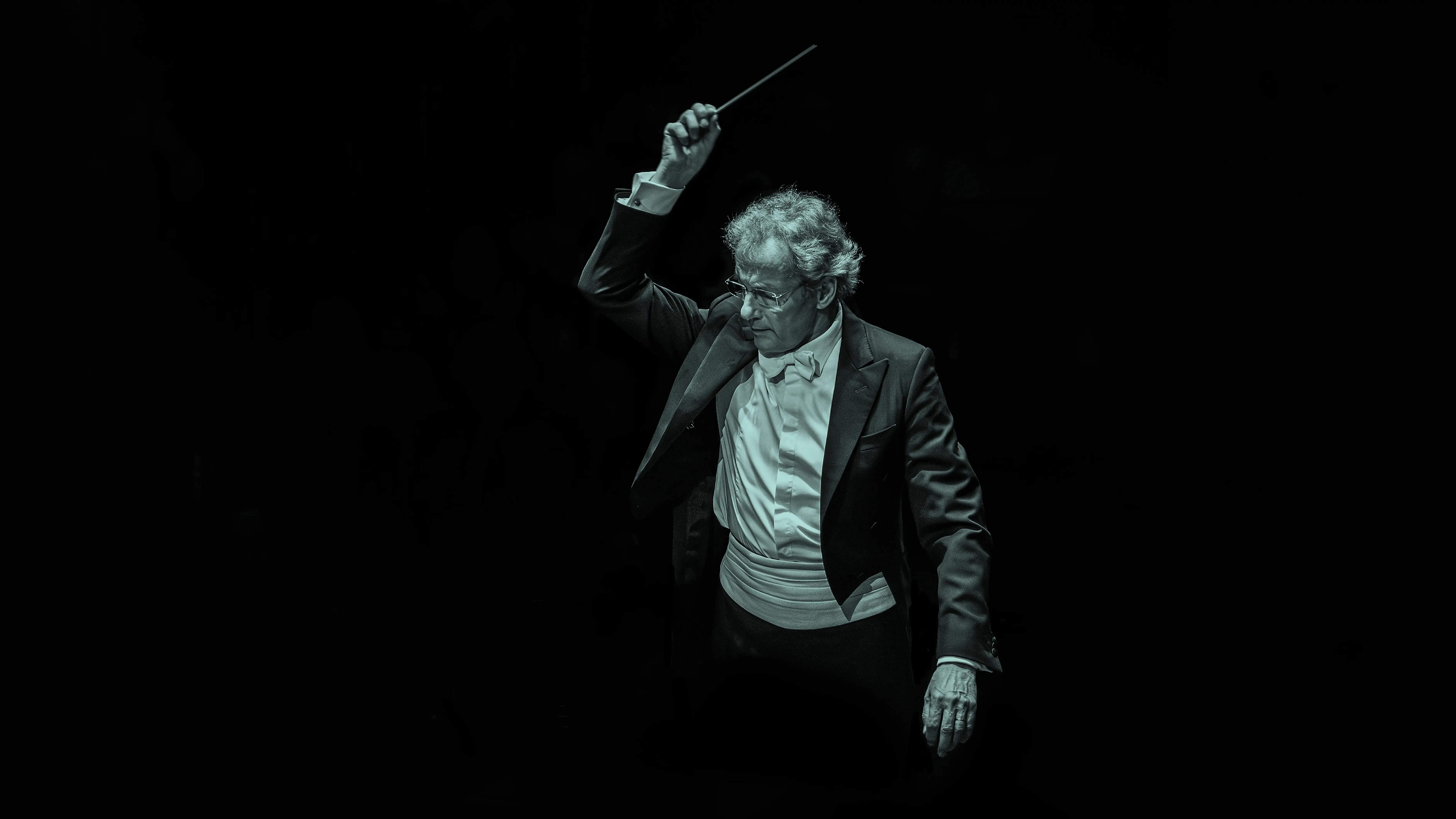 Franz Welser Möst in a tuxedo passionately leading an orchestra, baton raised, against a dark background.
