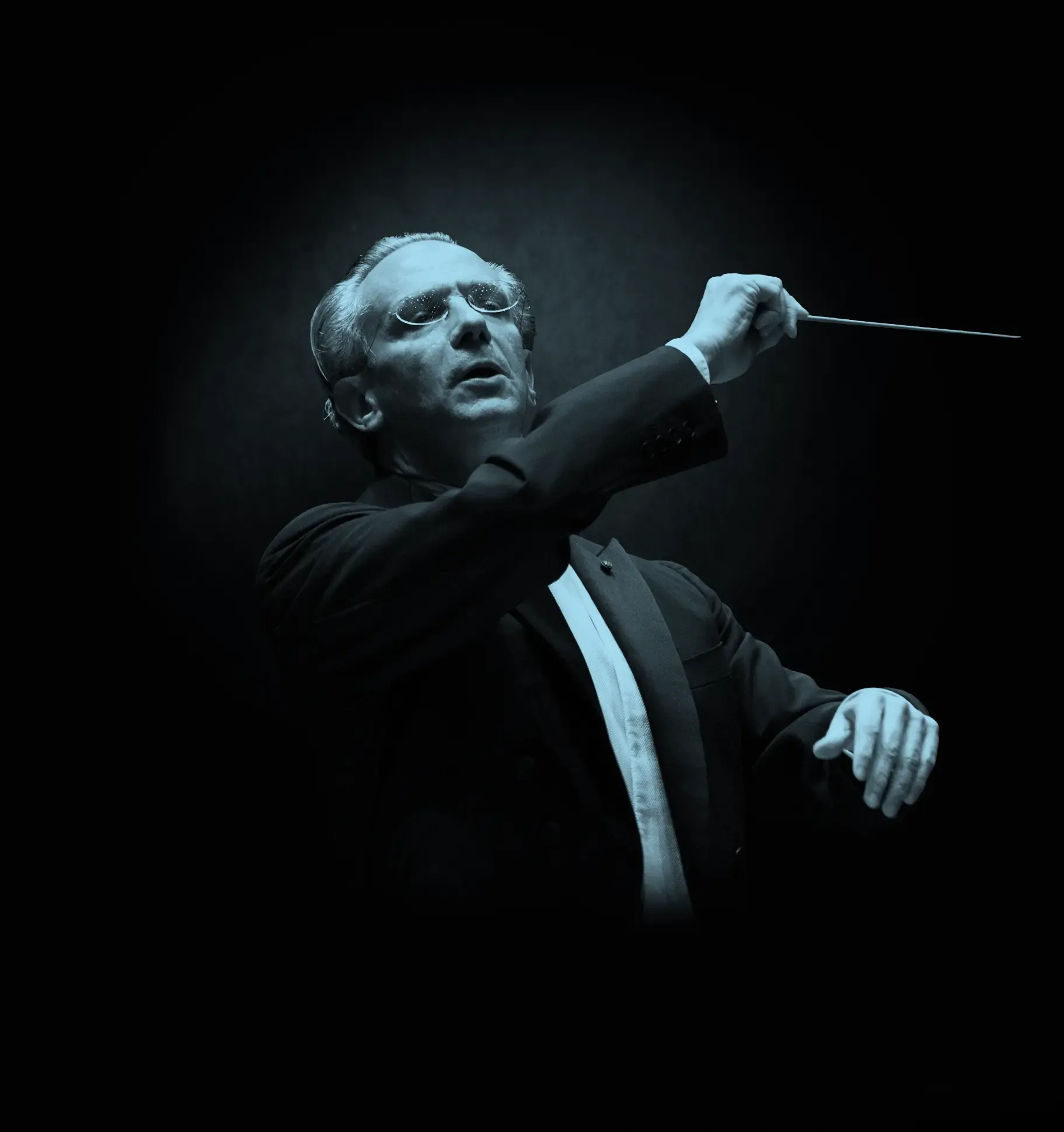 Conductor, Fabio Luisi, in a suit passionately leads an orchestra, holding a baton against a dark background.