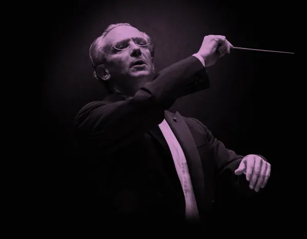 Conductor, Fabio Luisi, in a suit passionately leads an orchestra, holding a baton against a dark background.