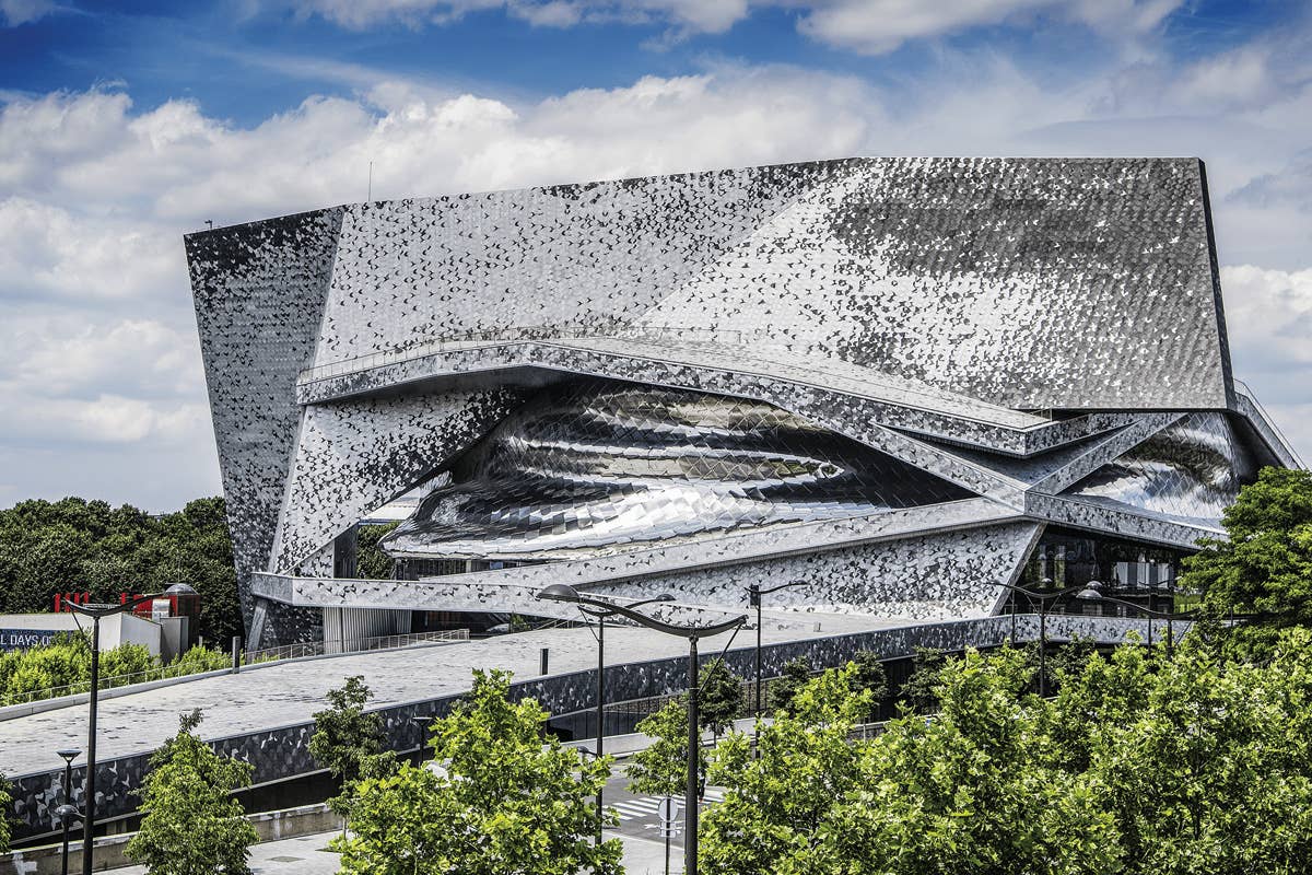 Modern architectural building with a metallic, angular facade under a partly cloudy sky, surrounded by green trees.