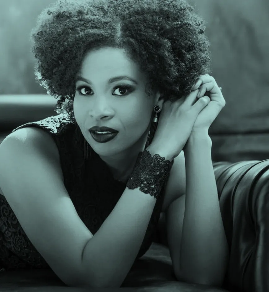Golda Schultz with curly hair and lace bracelet, resting on a sofa, looking directly at the camera with a calm expression.