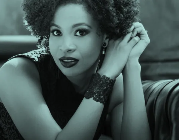 Golda Schultz with curly hair and lace bracelet, resting on a sofa, looking directly at the camera with a calm expression.