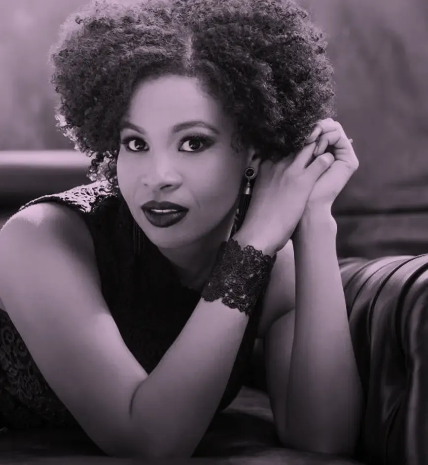 Golda Schultz with curly hair and lace bracelet, resting on a sofa, looking directly at the camera with a calm expression.