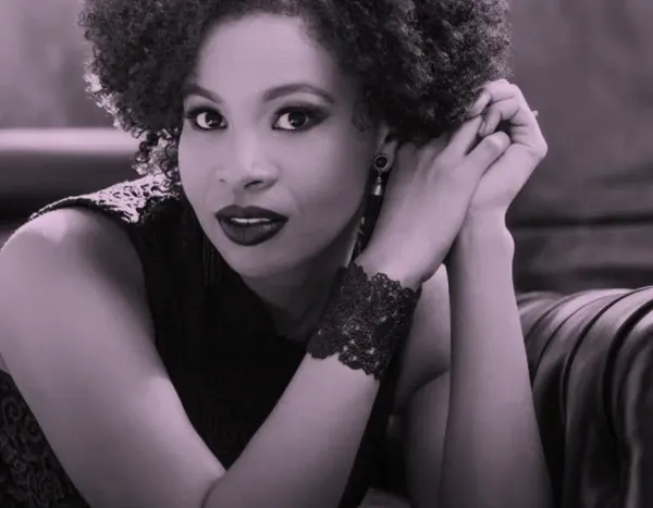 Golda Schultz with curly hair and lace bracelet, resting on a sofa, looking directly at the camera with a calm expression.
