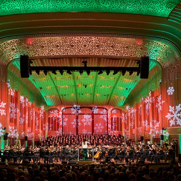 An orchestra sits on a stage bathed in green and red light with snowflake projections