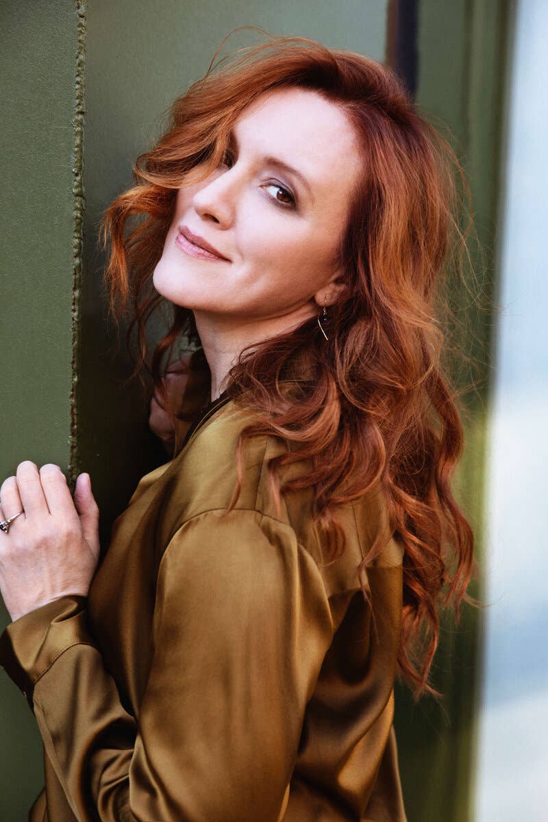 Sara Jakubiak with wavy red hair and a brown blouse leans against a green wall, smiling softly at the camera.