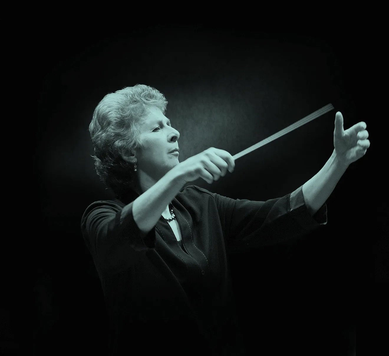British conductor, Jane Glover, leading an orchestra with a baton, focused expression, against a dark background.