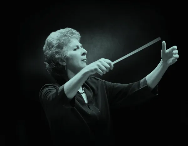 British conductor, Jane Glover, leading an orchestra with a baton, focused expression, against a dark background.