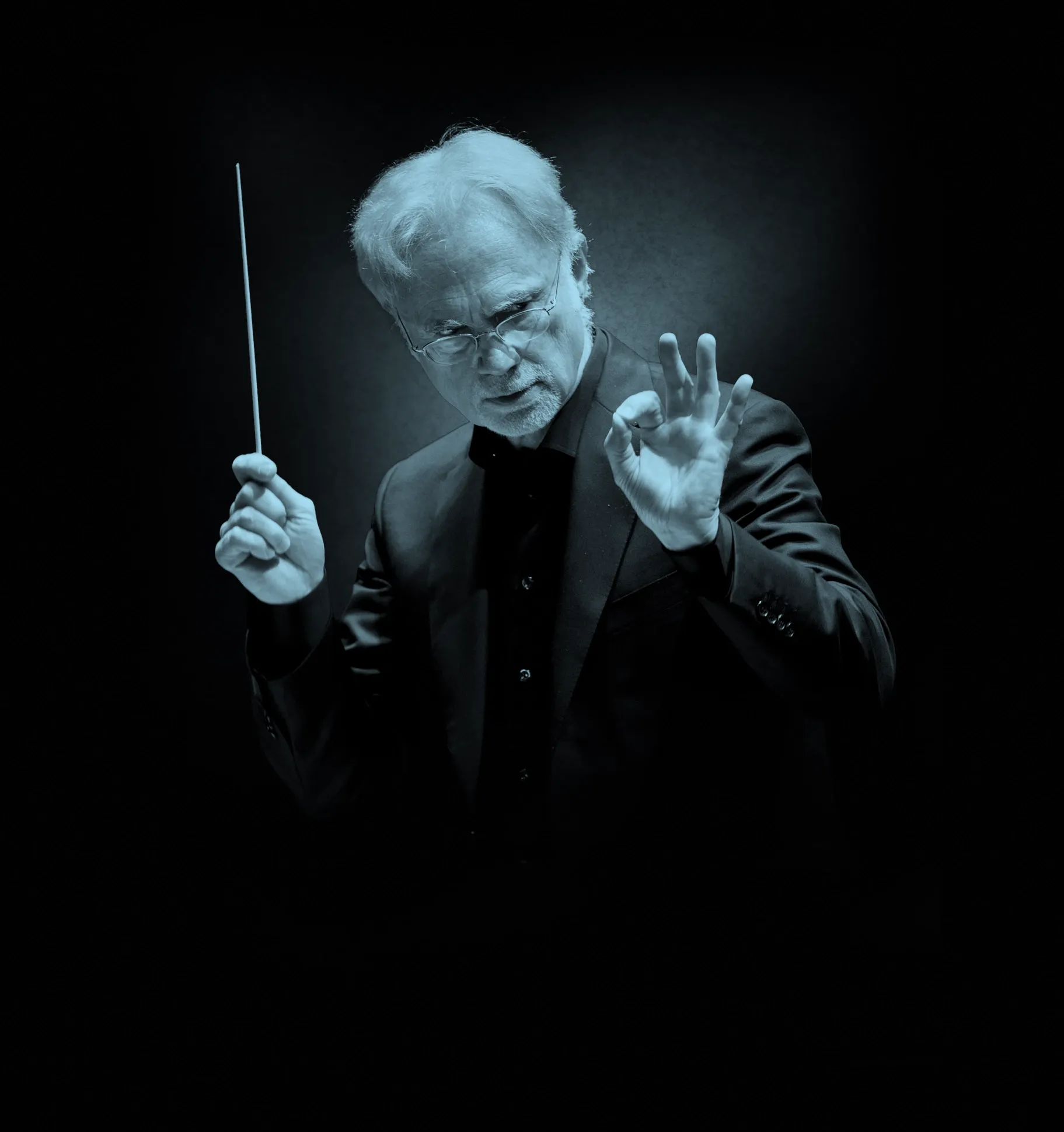 Conductor, John Adams, in a dark suit holding a baton, gesturing with his hand, set against a dark background.