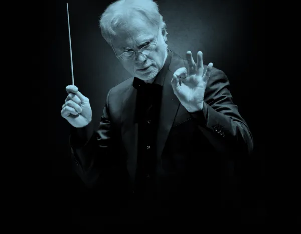 Conductor, John Adams, in a dark suit holding a baton, gesturing with his hand, set against a dark background.