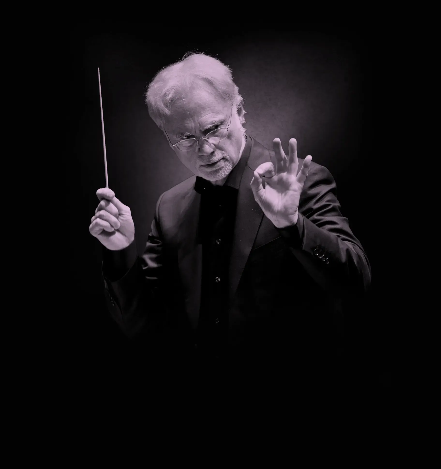Conductor, John Adams, in a dark suit holding a baton, gesturing with his hand, set against a dark background.
