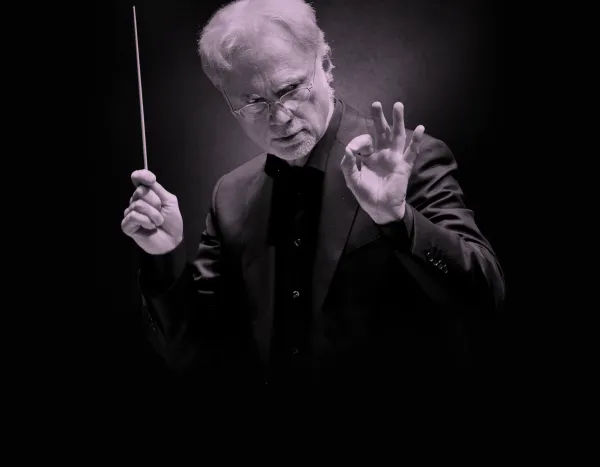 Conductor, John Adams, in a dark suit holding a baton, gesturing with his hand, set against a dark background.