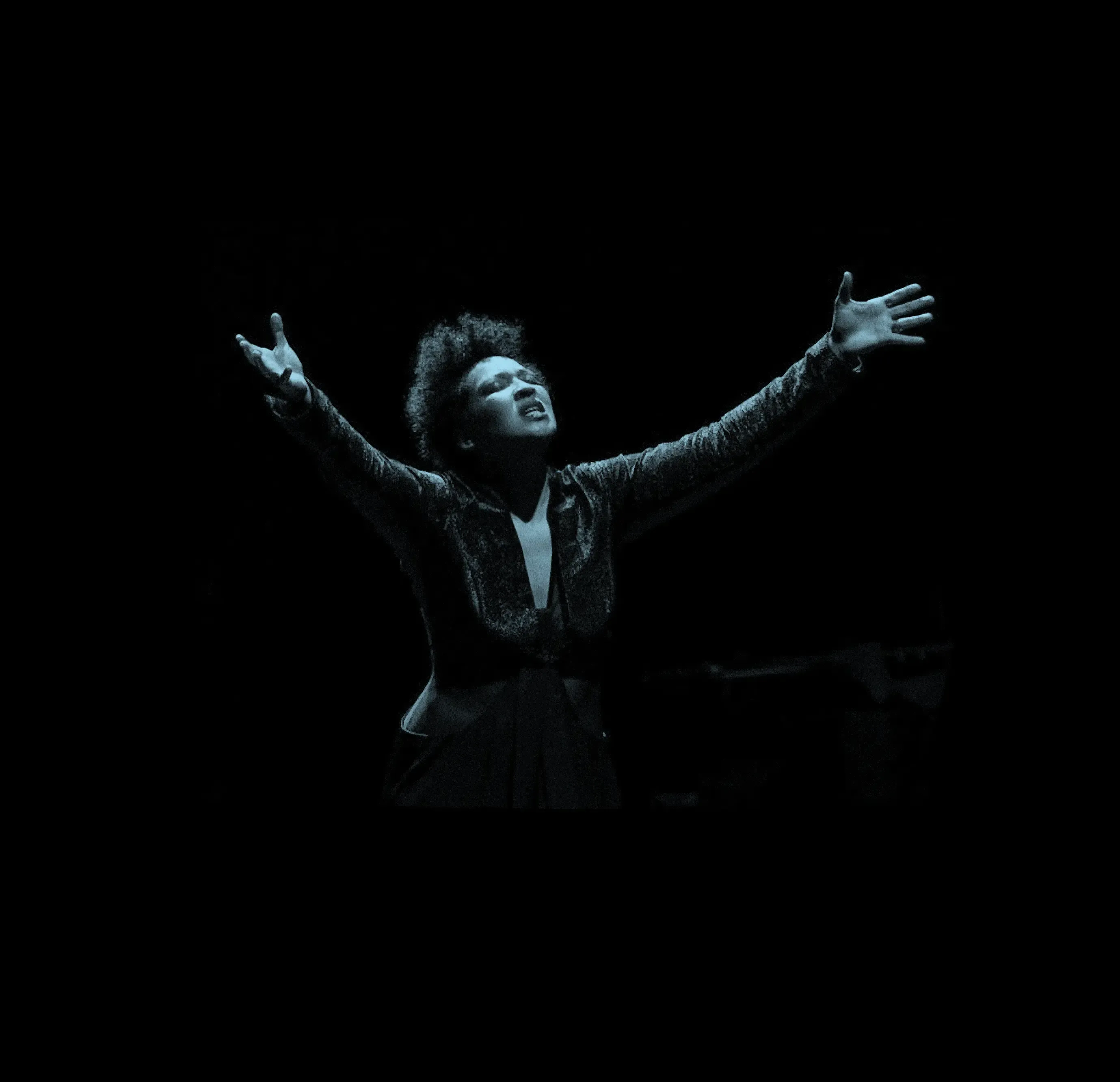 Julia Bullock with arms raised, passionately performing on stage in dramatic lighting.