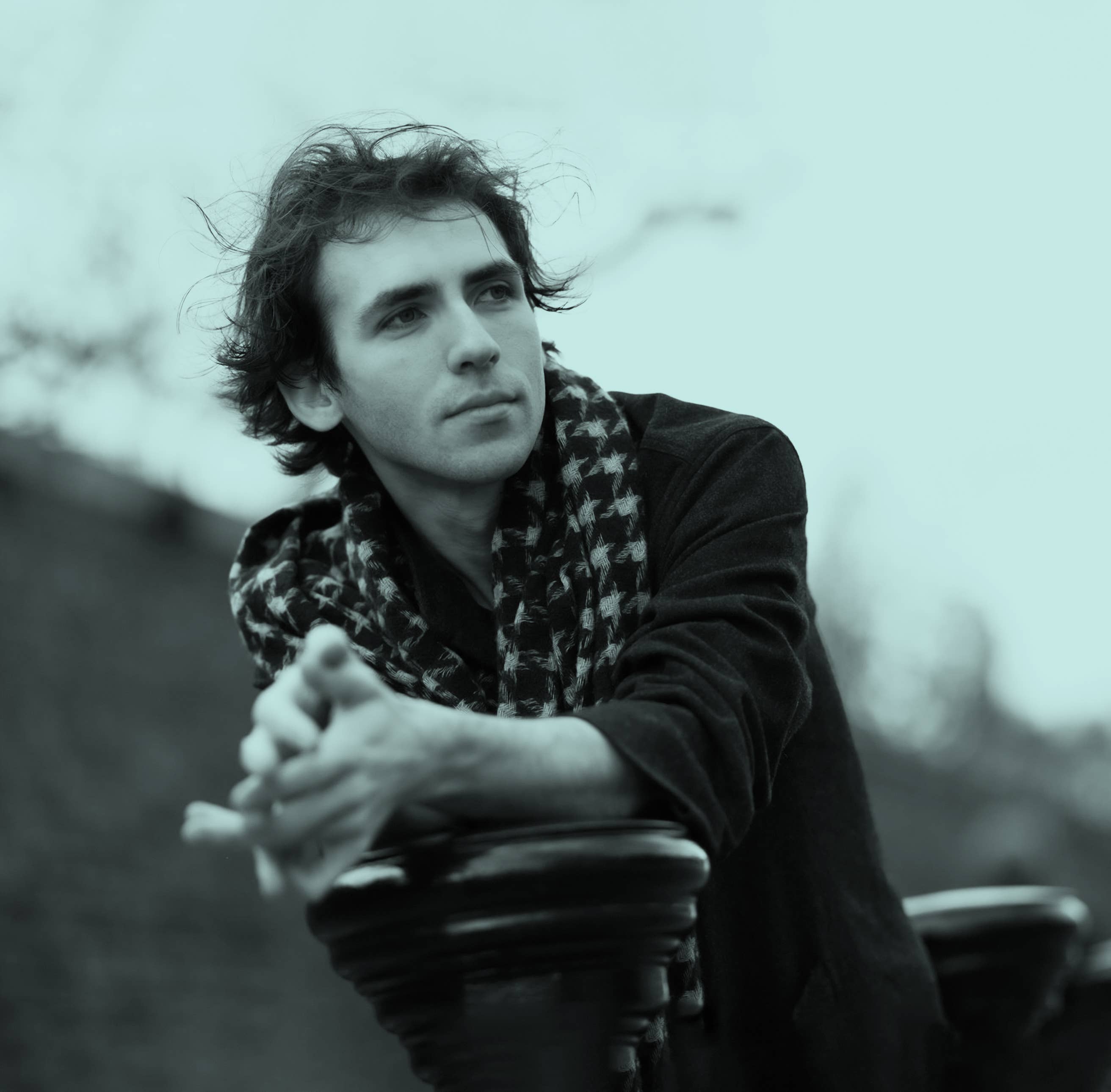 Alexandre Kantorow with tousled hair and scarf leans on a railing, gazing thoughtfully into the distance.
