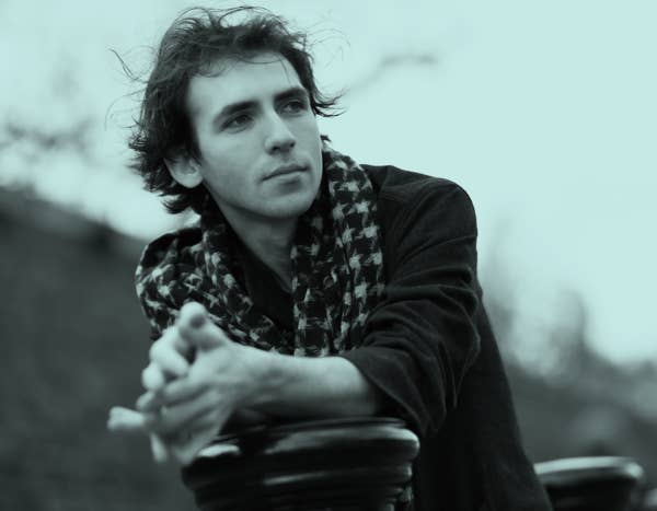 Alexandre Kantorow with tousled hair and scarf leans on a railing, gazing thoughtfully into the distance.