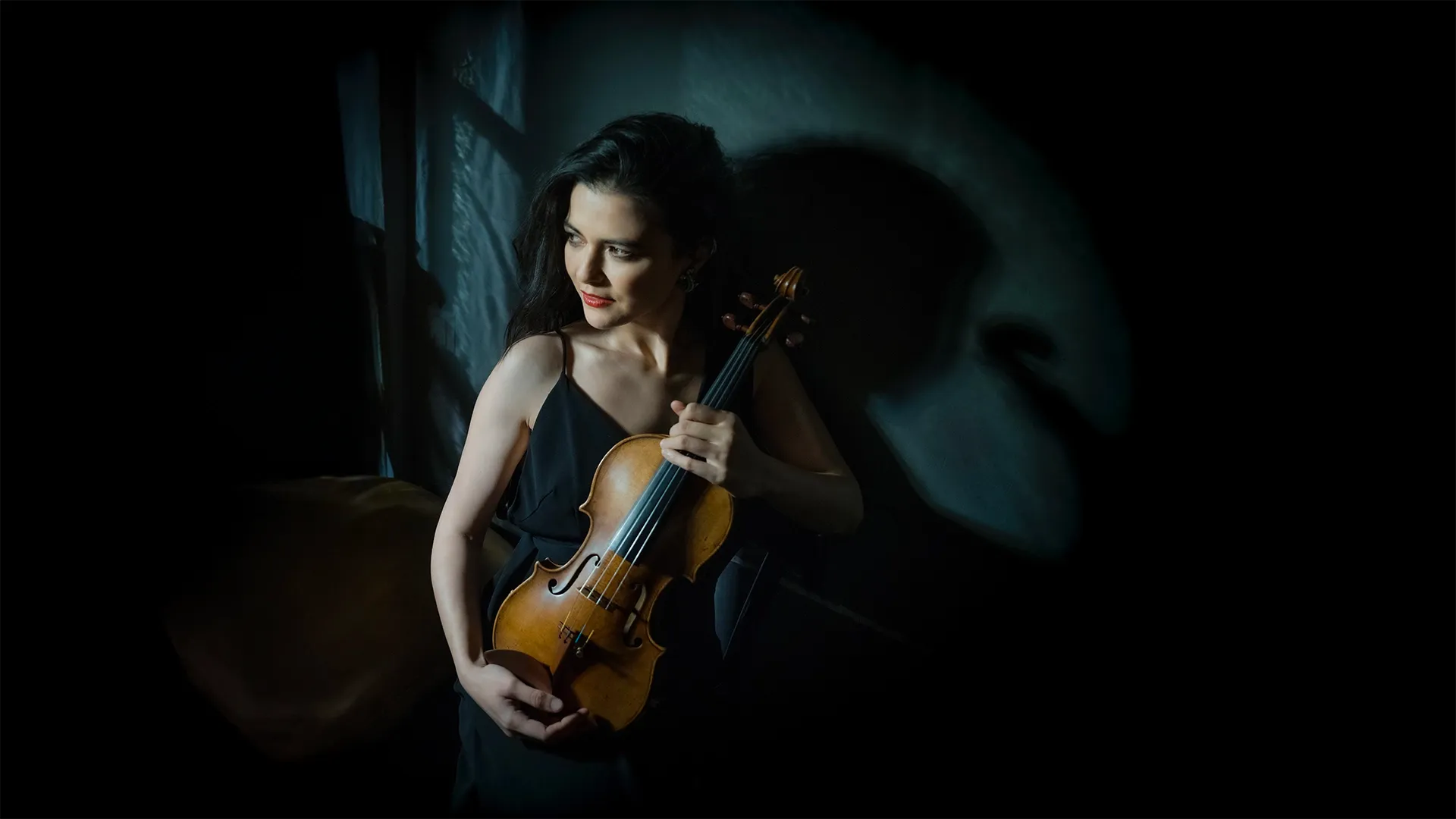 Woman in a dark dress holds a violin, standing in soft light with a shadowy background.