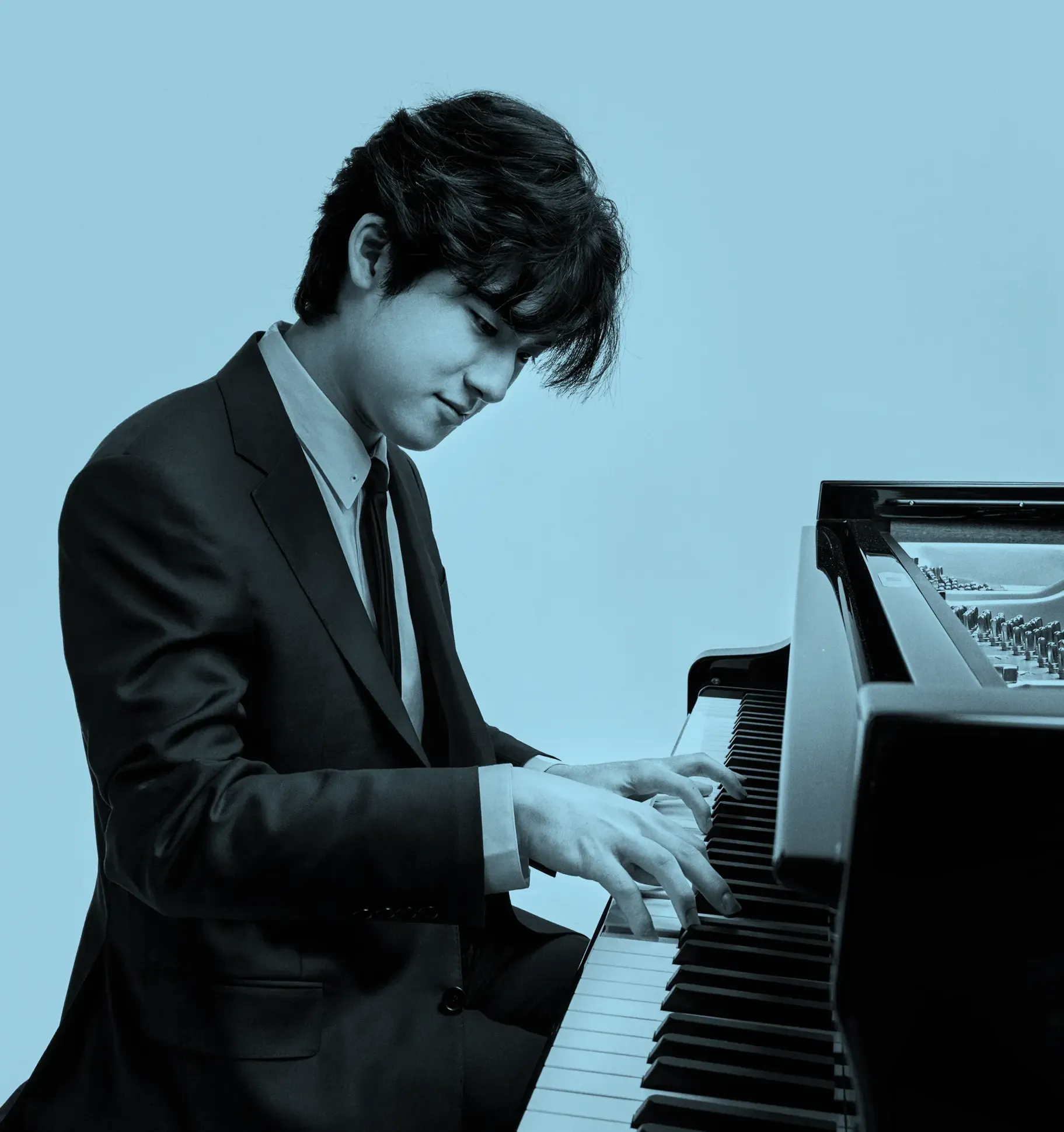 Yunchan Lim in a suit playing a grand piano with focus and concentration against a plain background.